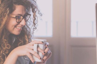 A woman smiling on her computer holding a white mug