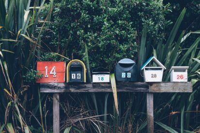 6 mailboxes in a row on a post with leaves and bushes in the background